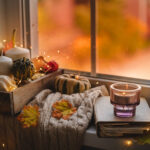 mini pumpkins and gourds on a white knit blanket, next to a book with a candle, showing a cozy setting